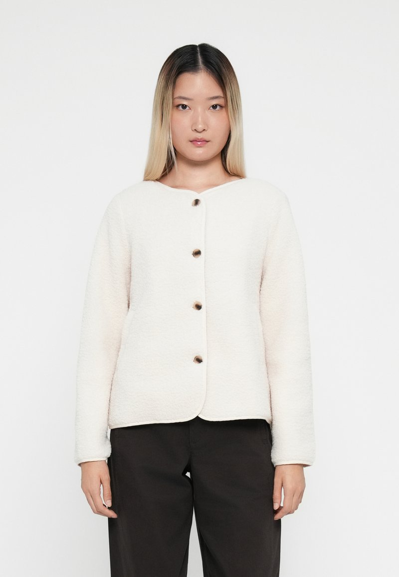 Veste en polaire crème avec un col arrondi, fermeture à boutons sur le devant et manches longues. Pantalon noir porté en dessous.