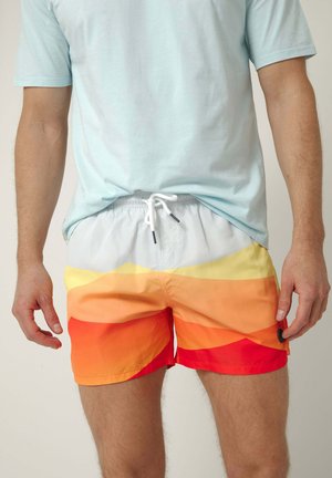 Hombre con camiseta azul claro y pantalones cortos de baño multicolor en naranja, amarillo y rojo con cordón blanco, de pie con los brazos relajados a los lados.