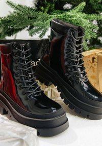 Czarne, błyszczące, sznurowane botki na platformie z grubymi, wytrzymałymi podeszwami, umieszczone na białej powierzchni, z zielonymi gałązkami jodły i owiniętymi prezentami w tle.