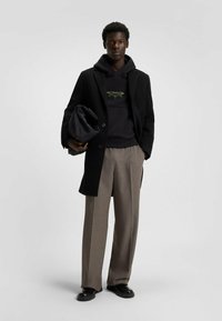 Cappotto di lana nera, pantaloni grigi e felpa nera con un logo verde. Tessuto liscio, vestibilità rilassata e silhouette oversize. Scarpe nere.
