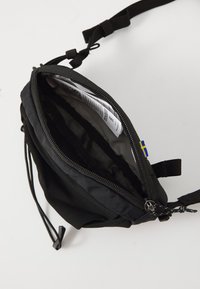 Borsa in tessuto nero con chiusura a zip, scomparto interno e tracolla regolabile. Presenta un piccolo etichetta con la bandiera svedese e foderatura interna testurizzata.