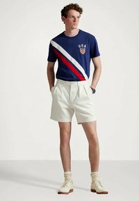 Mężczyzna w granatowej koszulce USA z czerwono-białymi diagonalnymi paskami, w białych szortach, beżowych skarpetkach i beżowych butach wizytowych, stoi z rękami w kieszeniach.