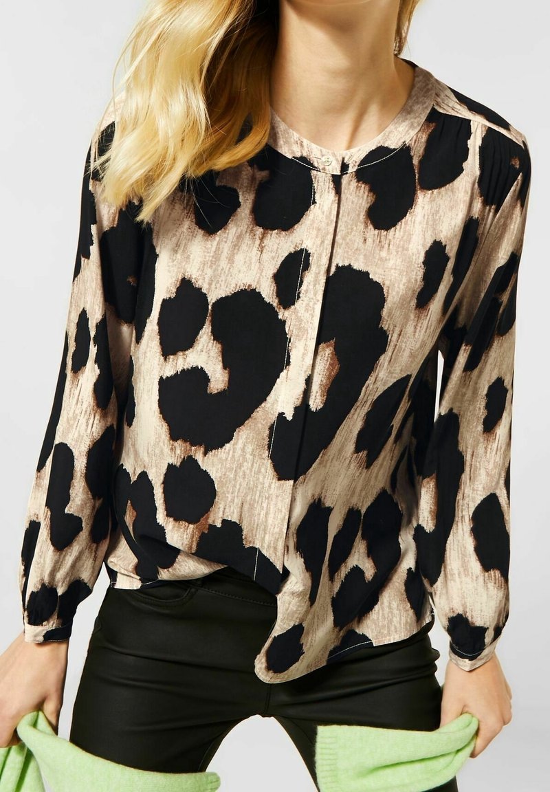 Femme portant un chemisier beige avec un grand imprimé animal abstrait noir et marron, associé à un pantalon noir et tenant un tissu vert.