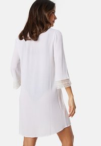 Abito bianco lungo fino al ginocchio con un tessuto testurizzato. Caratterizzato da maniche a tre quarti e dettagli in pizzo nei polsini.