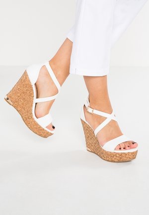 Mujer con sandalias de cuña blancas con correas y tacones de patrón de corcho, luciendo pantalones cropped blancos, contra un fondo blanco.