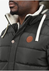 Gepolsterte schwarze Jacke mit weißem Lammfellkragen, verstellbarem Kordelzug und braunem Lederlogo-Badge auf der Vorderseite.