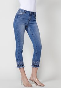 Jean capri en denim bleu clair avec un ourlet orné de motifs en diamants blancs et bleu foncé. Bords effilochés et design à cinq poches.