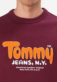 Hombre vestido con un sudadera burdeos con el texto texturizado en naranja "Tommy" y el texto en blanco "JEANS, N.Y. American Outdoor Original New York, NY, U.S.A."