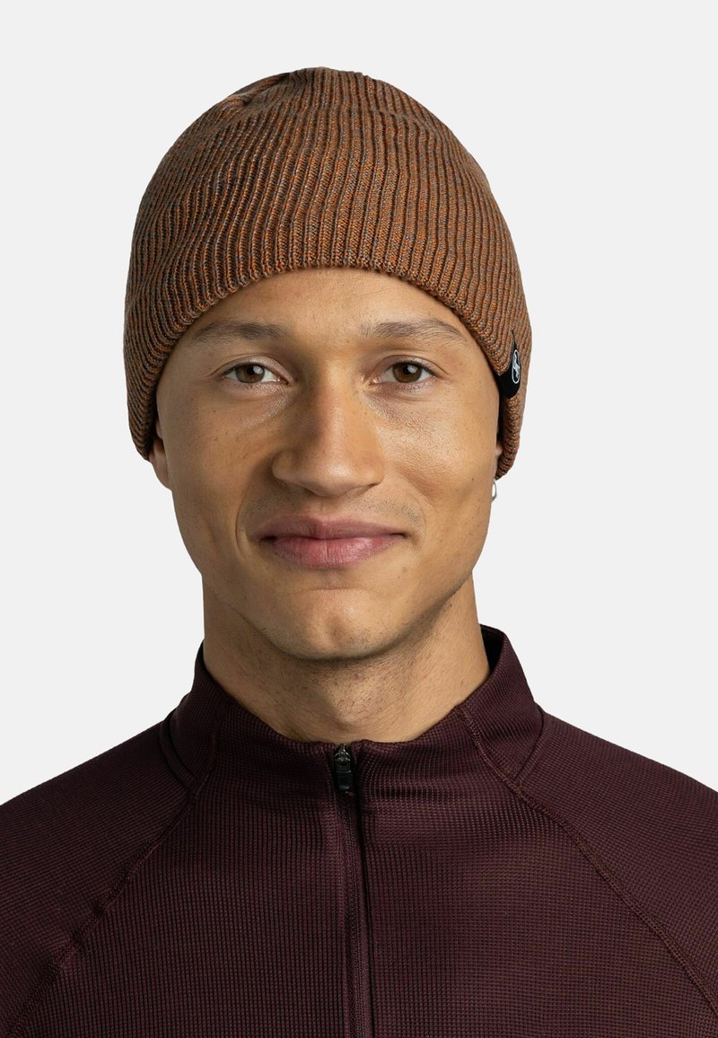 Brown knit beanie with a ribbed texture, featuring a small black label accent. Worn by a person in a dark maroon zip-up shirt.