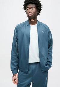 Μπλε αθλητικό μπουφάν με λείο φινίρισμα, που διαθέτει λευκές ρίγες και λογότυπο στο στήθος, συνδυασμένο με αντίστοιχο παντελόνι και ανοιχτόχρωμο πουκάμισο.