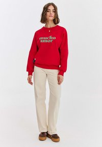 Czerwona bluza z napisem "mucho amor" w kolorze zielonym, z okrągłym dekoltem i szerokimi rękawami. Połączona z kremowymi spodniami o szerokim kroju i brązowymi butami.