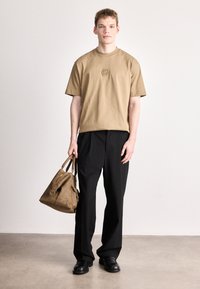 Μπεζ βαμβακερό t-shirt με ανάγλυφο λογότυπο, σε συνδυασμό με μαύρο παντελόνι φαρδιάς γραμμής. Το μοντέλο κρατάει καφέ δερμάτινη τσάντα με κοντράστ μεταλλικά στοιχεία.