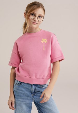 Giovane donna con occhiali che indossa una felpa rosa a maniche corte con un ricamo di palma gialla e jeans blu su uno sfondo neutro.