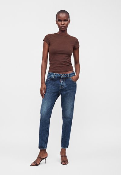 Femme portant un haut marron moulant à manches courtes, un jean bleu taille haute et des sandales marron à talons, debout devant un fond clair uni.