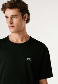 Camiseta negra de algodón con cuello redondo y mangas cortas. Pequeño logo blanco "PJL" en el lado izquierdo del pecho. Textura suave.