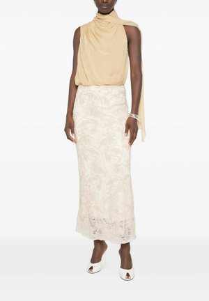 Vrouw draagt een beige mouwloze drapering top met sjaaldetail, witte geborduurde kanten rok, zilveren armband en witte open-teenhakken.
