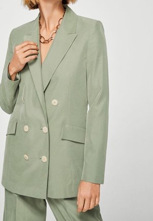 Woman wearing a sage green double-breasted blazer with beige buttons and matching pants, accessorized with a chunky gold chain necklace.