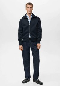 Giacca con cerniera blu navy dalla texture morbida, abbinata a camicia azzurro chiaro, pantaloni in denim scuro e scarpe in pelle nere. Design semplice e moderno.