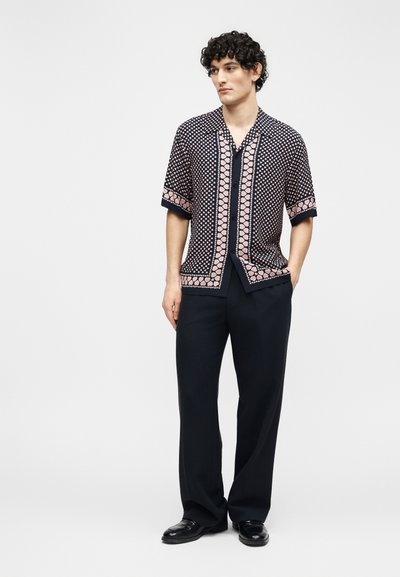 Άνδρας με κατσαρά μαλλιά που φοράει μια κοντομάνικη πουκάμισο με μαύρο και ροζ σχέδιο, μαύρο παντελόνι και μαύρα παπούτσια, στέκεται με το ένα χέρι στην τσέπη.
