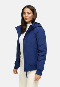 Marineblauwe hooded jack met ritssluiting, geribbelde manchetten en een zachte textuur. Het heeft een logo op de mouw en een binnenvoering.