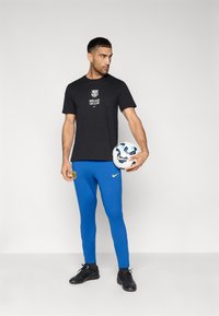 Μαύρο βαμβακερό μπλουζάκι με λογότυπο και κείμενο, μπλε φόρμα με έμβλημα συλλόγου και μαύρα αθλητικά παπούτσια. Κρατάει μια μπάλα ποδοσφαίρου.