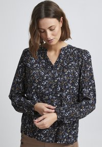 Blusa floreale in navy con accenti bianchi e dorati. Presenta un collo a V, maniche lunghe con polsini arricciati e un tessuto testurizzato.