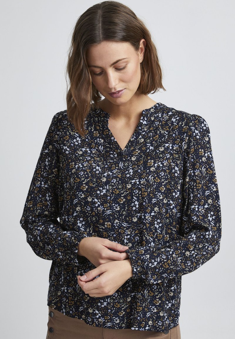 Blusa floreale in navy con accenti bianchi e dorati. Presenta un collo a V, maniche lunghe con polsini arricciati e un tessuto testurizzato.
