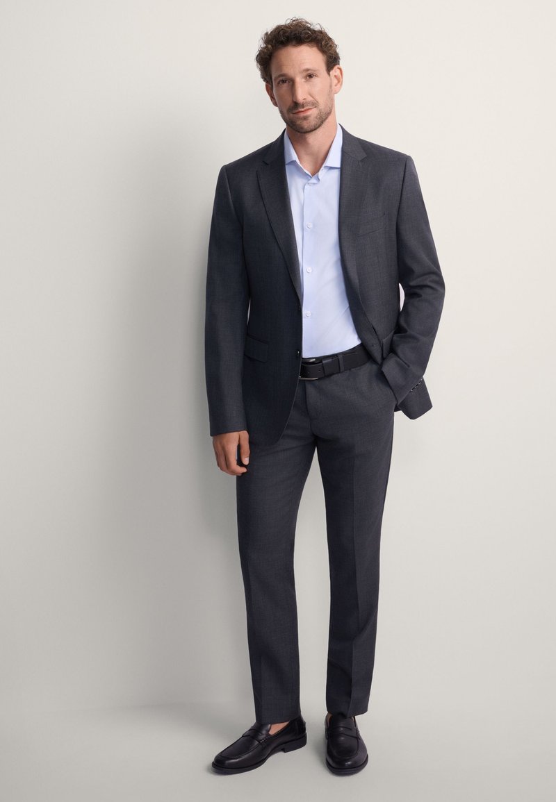 Ciemnoszary garnitur z jasnoniebieską koszulą. Dopasowany krój, marynarka na jeden guzik, spodnie z prostymi nogawkami oraz czarne wsuwane buty.