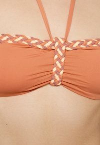 Top de bikini semelhante a um soutien em tecido pêssego, com um detalhe trançado em laranja claro, taupe e creme ao centro, apresentando alças ajustáveis.