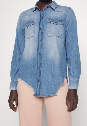 Person iført en lyseblå denim skjorte med knapper, frynset kant og to brystlommer, kombineret med lyserøde bukser.