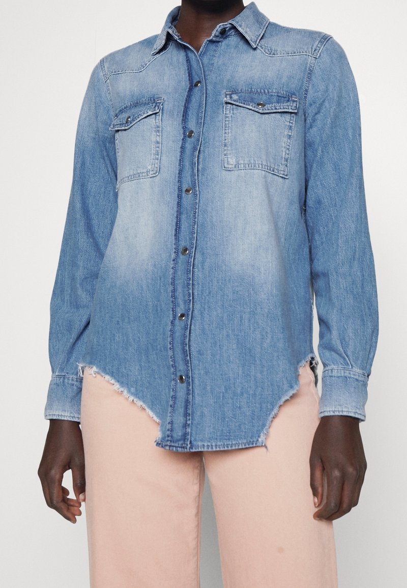 Personne portant une chemise en denim bleu clair à boutons, avec un ourlet effiloché et deux poches poitrine, associée à un pantalon rose clair.