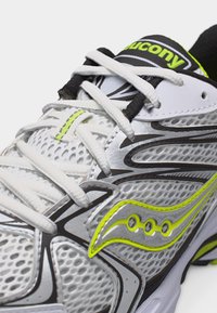 Zapatilla de running Saucony blanca y gris con detalles en amarillo neón y cordones blancos sobre un fondo claro, mostrando detalles de la parte superior y el logo.