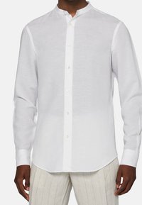 Camicia bianca a maniche lunghe realizzata in tessuto leggero. Presenta un colletto alla coreana, una patta frontale con bottoni e una vestibilità comoda. Superficie strutturata.