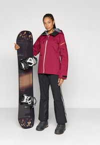 Mørkerød snejakke med lynlås foran og grå hætte, kombineret med sorte snebukser med hvide striber og sorte snowboardstøvler. Et snowboard med bindinger holdes.