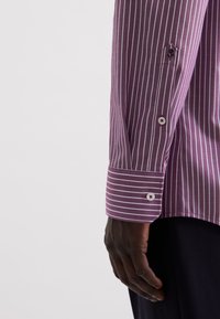 Dunkelhäutige Hand neben einem lila-weißen, senkrecht gestreiften Hemdsärmel mit geknöpftem Manschettenverschluss vor einem einfarbigen, hellen Hintergrund.