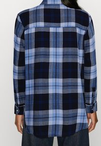 Vue de dos d'une personne portant une chemise à manches longues à carreaux bleu et noir avec des poignets boutonnés et un pantalon foncé.