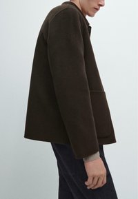 Giacca marrone, dalla texture ruvida e dal Taglio morbido, con colletto e tasche laterali, abbinata a jeans scuri e dettagli a coste sui polsini.