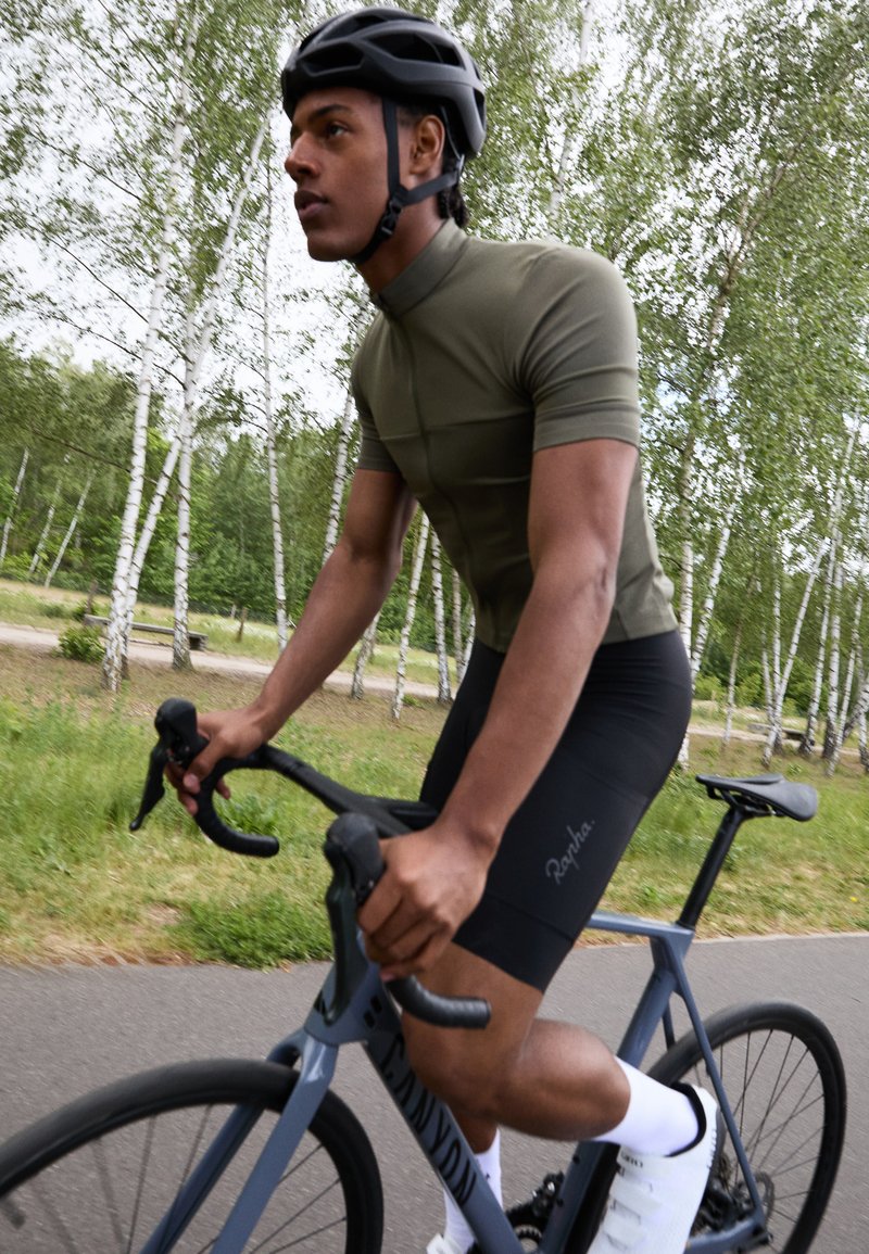 Sportiska riteņbraukšanas tērps, kas sastāv no pieguļošas olīvzaļas īsām piedurknēm un melniem šortiem. Riteņbraucējs valkā melnu ķiveri un baltas zeķes, turēdams pie stūres.