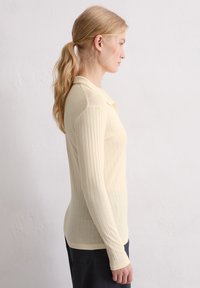 Frau mit blonden Haaren im Pferdeschwanz, die ein cremefarbenes, geripptes Langarm-Polohemd und dunkle Hose trägt, steht im Profil vor einer schlichten Wand.