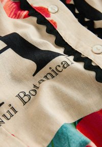 Gros plan sur un tissu beige avec un galon ondulé noir, des boutons blancs et un texte noir indiquant « Nui Botanical », avec des accents d'imprimé floral rouge et vert.