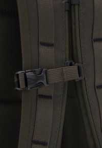 Détail d'un sac à dos vert olive montrant une boucle en plastique noir, des sangles ajustables et un tissu texturé avec un motif en grille et des coutures renforcées.
