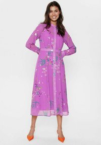 Rochie transparentă, cu mâneci lungi și butoni, de culoare violet intens, cu modele florale, talie strânsă și tocuri portocalii contrastante.