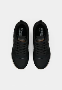 Um par de ténis Skechers pretos com atacadores, detalhes em logotipo castanho e palmilhas em espuma memória, vistos de cima sobre um fundo branco.