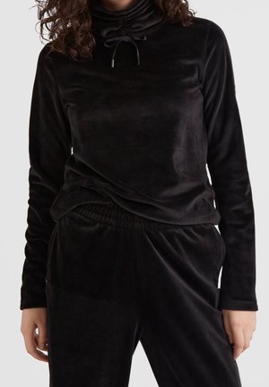 Persoon draagt een zwart fluweelachtig langewerpshirt met een coltrui en trekkoord, gecombineerd met bijpassende zwarte fluweelachtige broek.