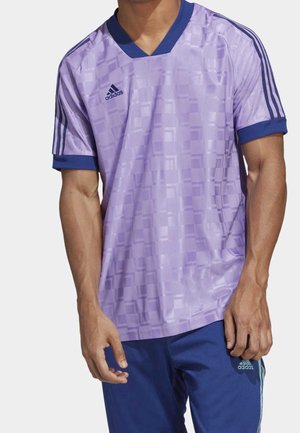 Mannelijk model draagt een lichtpaars Adidas-sportshirt met donkerblauwe accenten en bijpassende donkerblauwe Adidas-broek.