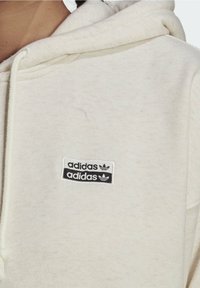 Detailní záběr na krémovou mikinu s kapucí, na které jsou na hrudi umístěny dva logo nášivky Adidas v černé a bílé barvě, s detaily šňůrek u kapuce.