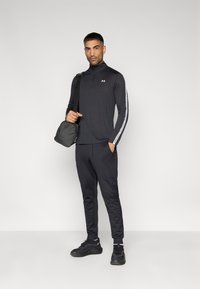 Μαύρο αθλητικό σύνολο που περιλαμβάνει μακρυμάνικο πουκάμισο με ψεύτικο γιακά και λευκές ρίγες, καθώς και αντίστοιχο παντελόνι φόρμας, συνδυασμένο με μαύρα αθλητικά παπούτσια και τσάντα.