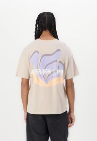 Beige bomull t-skjorte med et stort abstrakt design i lys lilla og oransje på baksiden, med teksten "ATELIER 199" under.