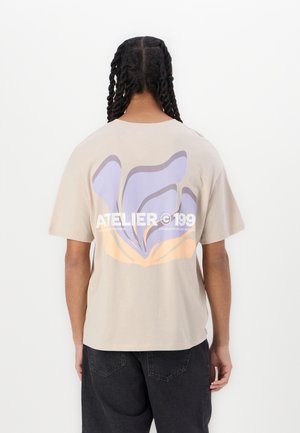 Beige katoenen t-shirt met een groot abstract ontwerp in lichtpaars en oranje op de achterkant, met de tekst "ATELIER 199" daaronder.