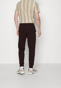 Braune Jogginghosen mit Bündchen, elastischem Bund und seitlichen Taschen; kombiniert mit einem beigen, gestreiften Kurzarmhemd und hellen Sneakers.
